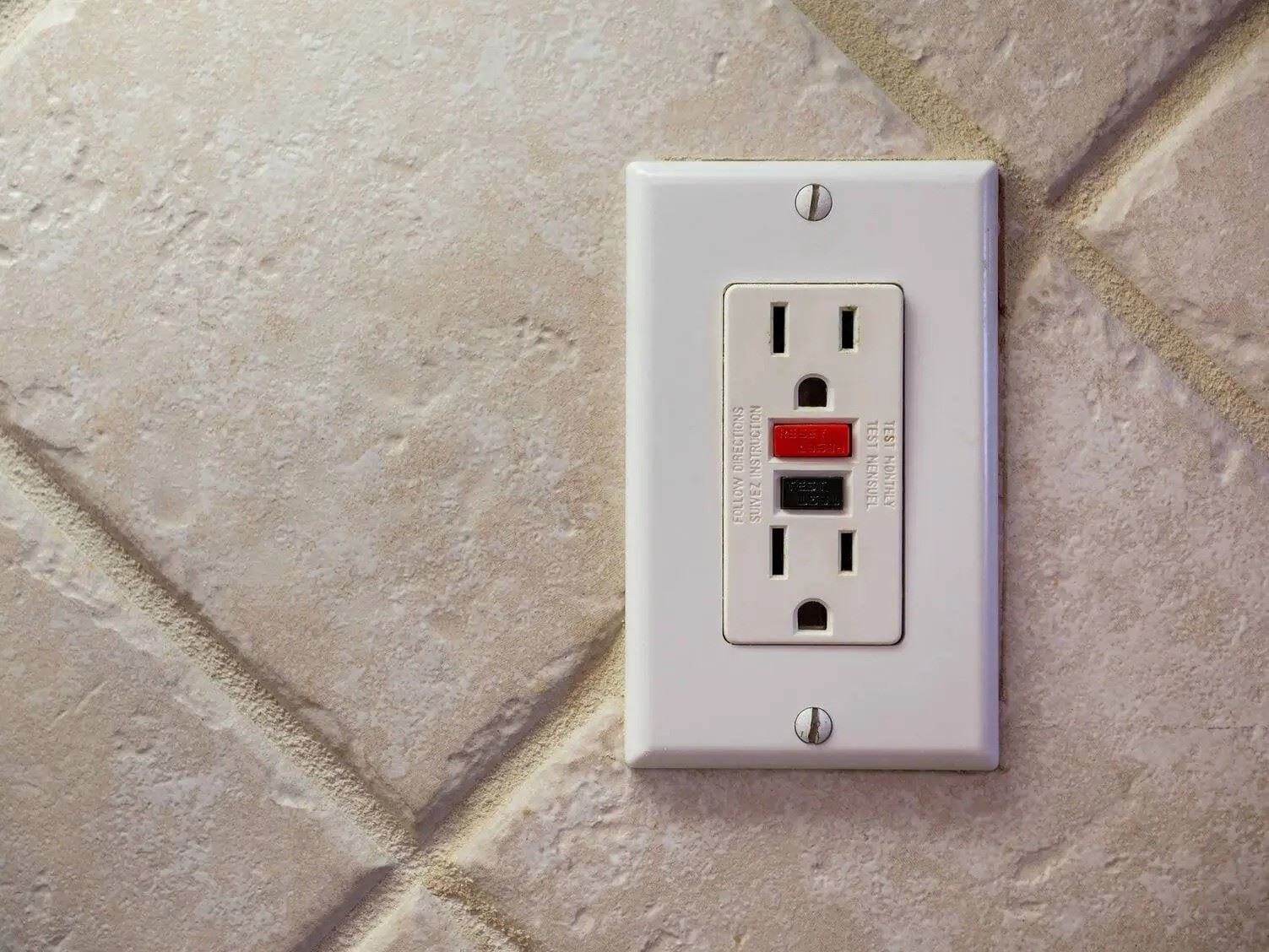 A GFCI (Ground Fault Circuit Interrupter) electrical outlet installed on a tiled wall, featuring two vertical plug slots, a red reset button, and a black test button.