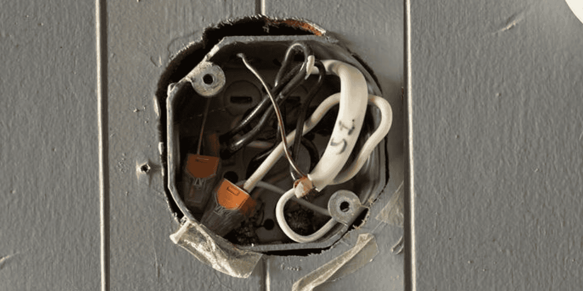 An open electrical junction box recessed into a gray paneled wall, showing a cluster of black, white, and bare copper wires. Several connections are secured with bright orange WAGO lever-nut connectors, which are clearly visible in the lower left portion of the box. The surrounding wall paint is chipped, and a piece of frayed white tape is partially attached to the metal box.          Ask ChatGPT